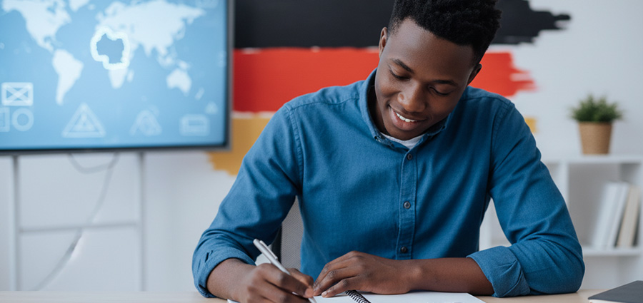Happy-kenyan-young-man-studying-german-in-a-class-with-german-flag-in-the-background.-german-language-course-for-work-abroad.
